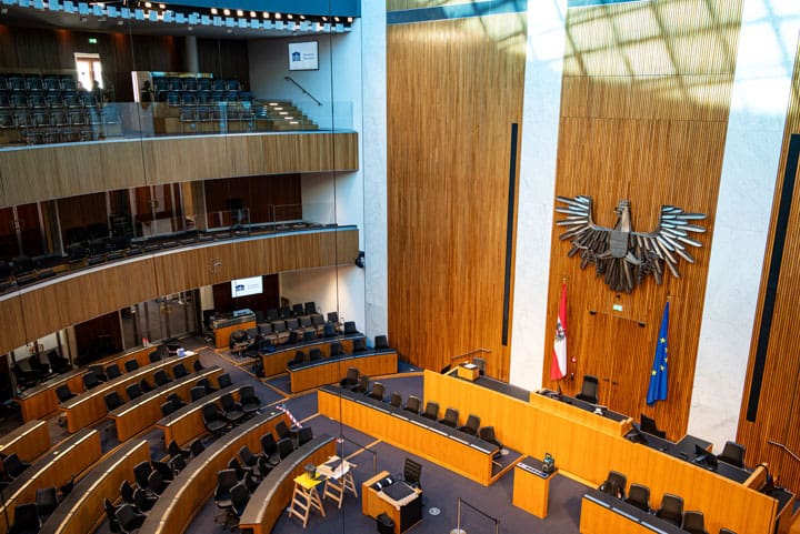 Geräumiger Plenarsaal mit Holzvertäfelung, leeren Rängen, großem Emblem, Fahnen und Rednerpult – symbolisiert formelle parlamentarische Umgebung bei Falschaussagen im U-Ausschuss