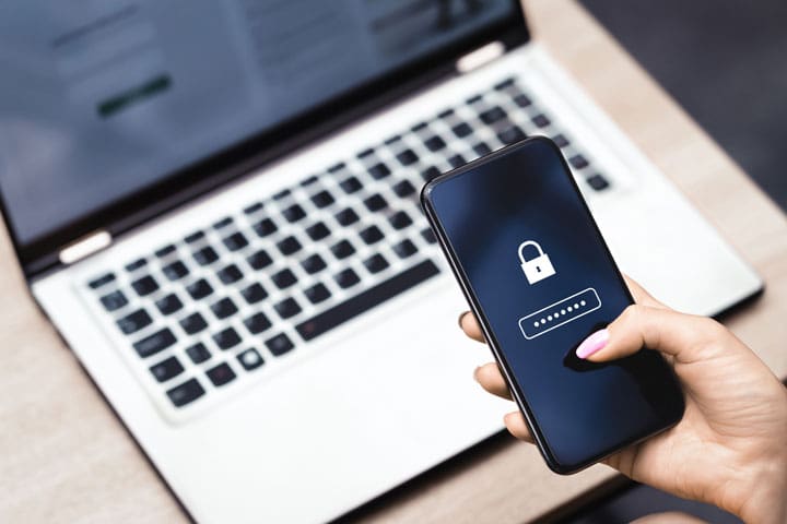 Person hält Smartphone mit Schloss-Symbol und Passwortfeld vor offenem Laptop – symbolisiert Sicherheit und Schutz von Daten bei Hausdurchsuchungen