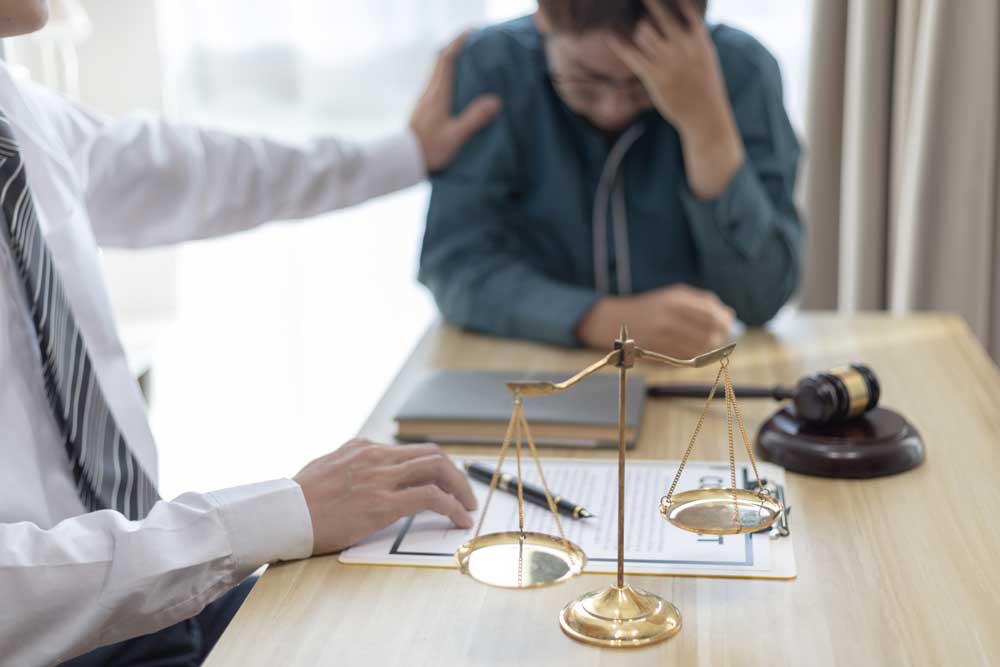 Ein Anwalt im Anzug tröstet einen verzweifelten Mandanten an einem Tisch. Eine Justizwaage und ein Richterhammer stehen im Fokus und symbolisieren rechtliche Beratung und Unterstützung.